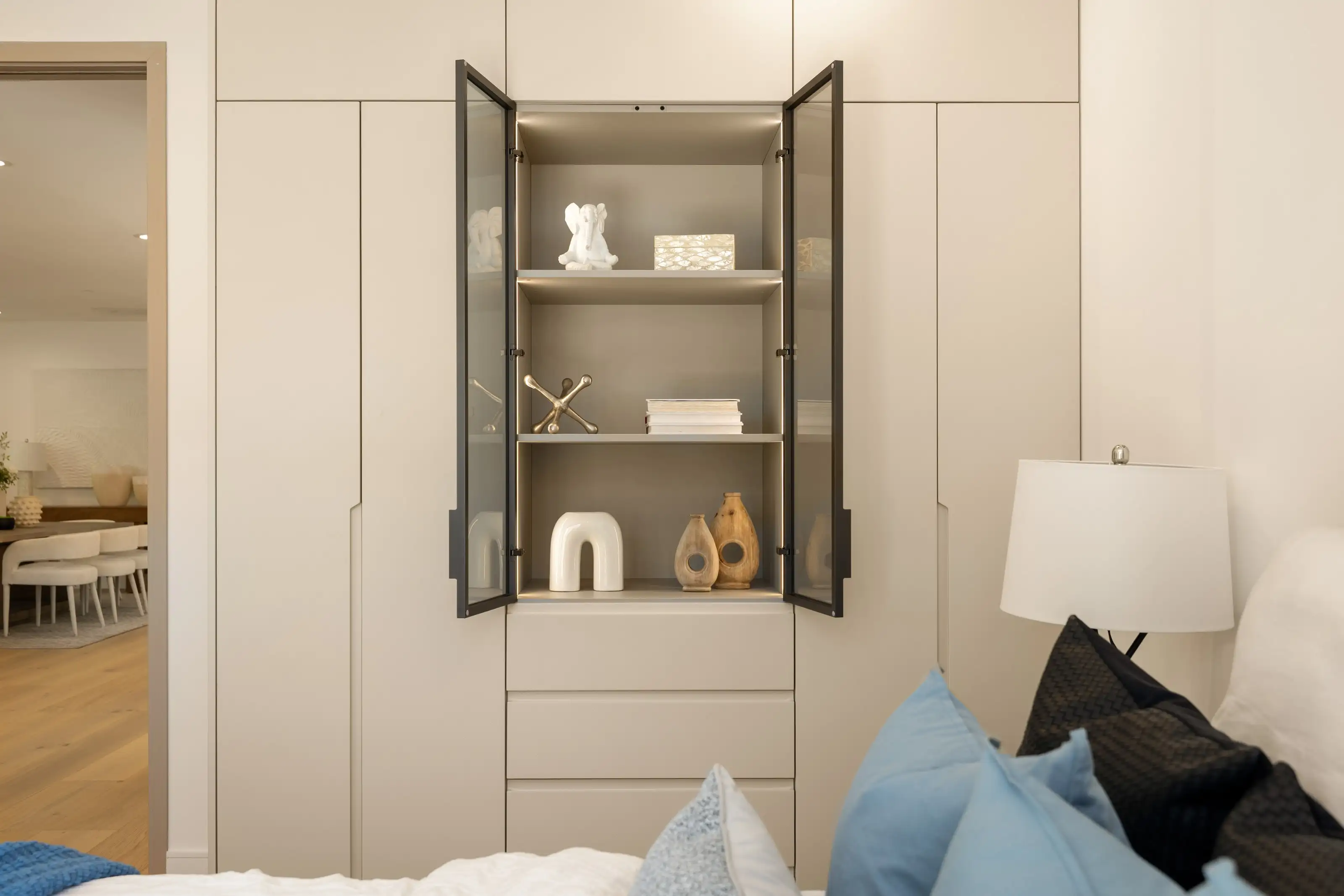 Floor to ceiling handleless bedroom wardrobes in warm greige with a lit glass front display cabinet and drawers below