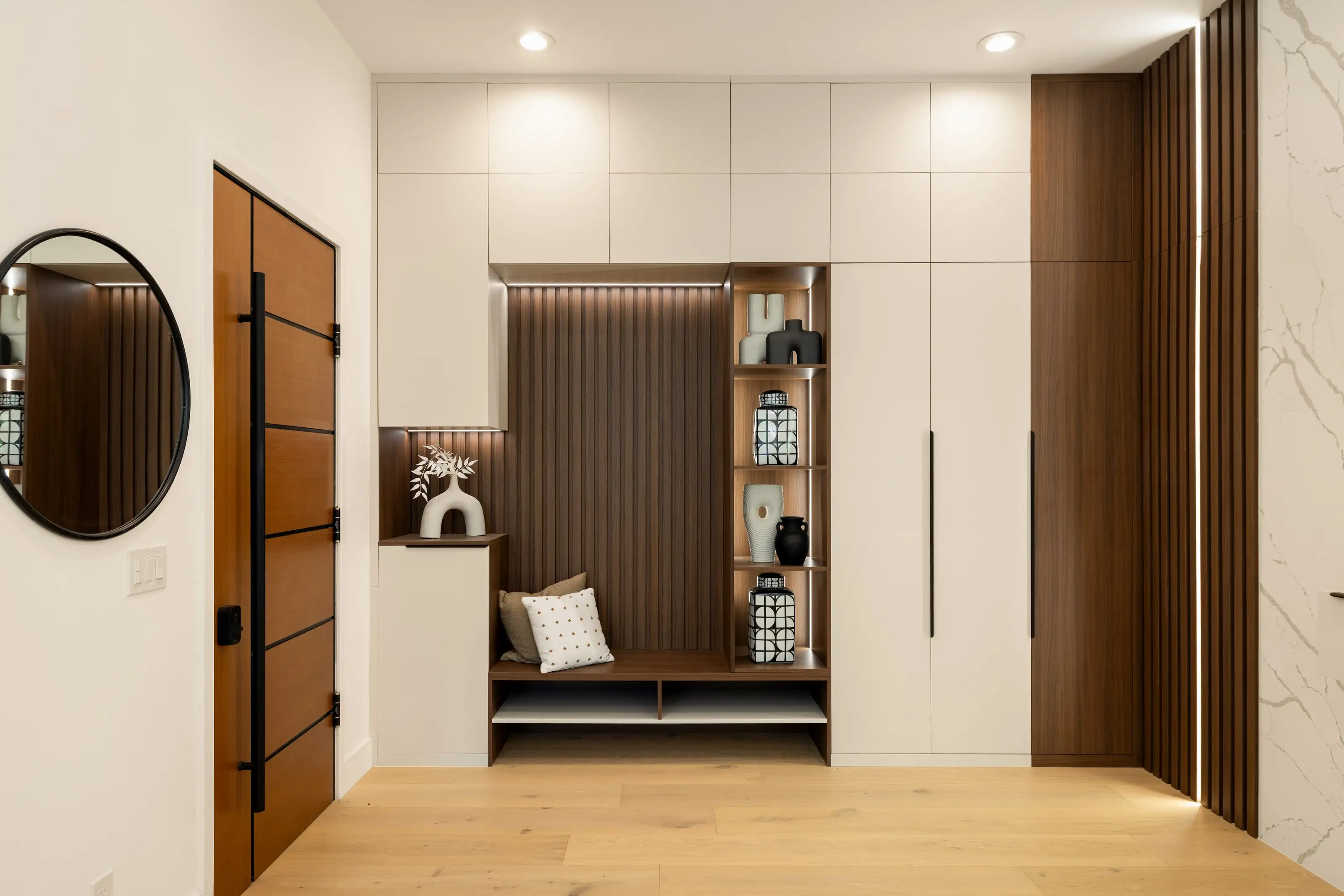 Entryway storage wall with handleless white cabinetry, walnut fluted back panel, bench seat and lit display niches