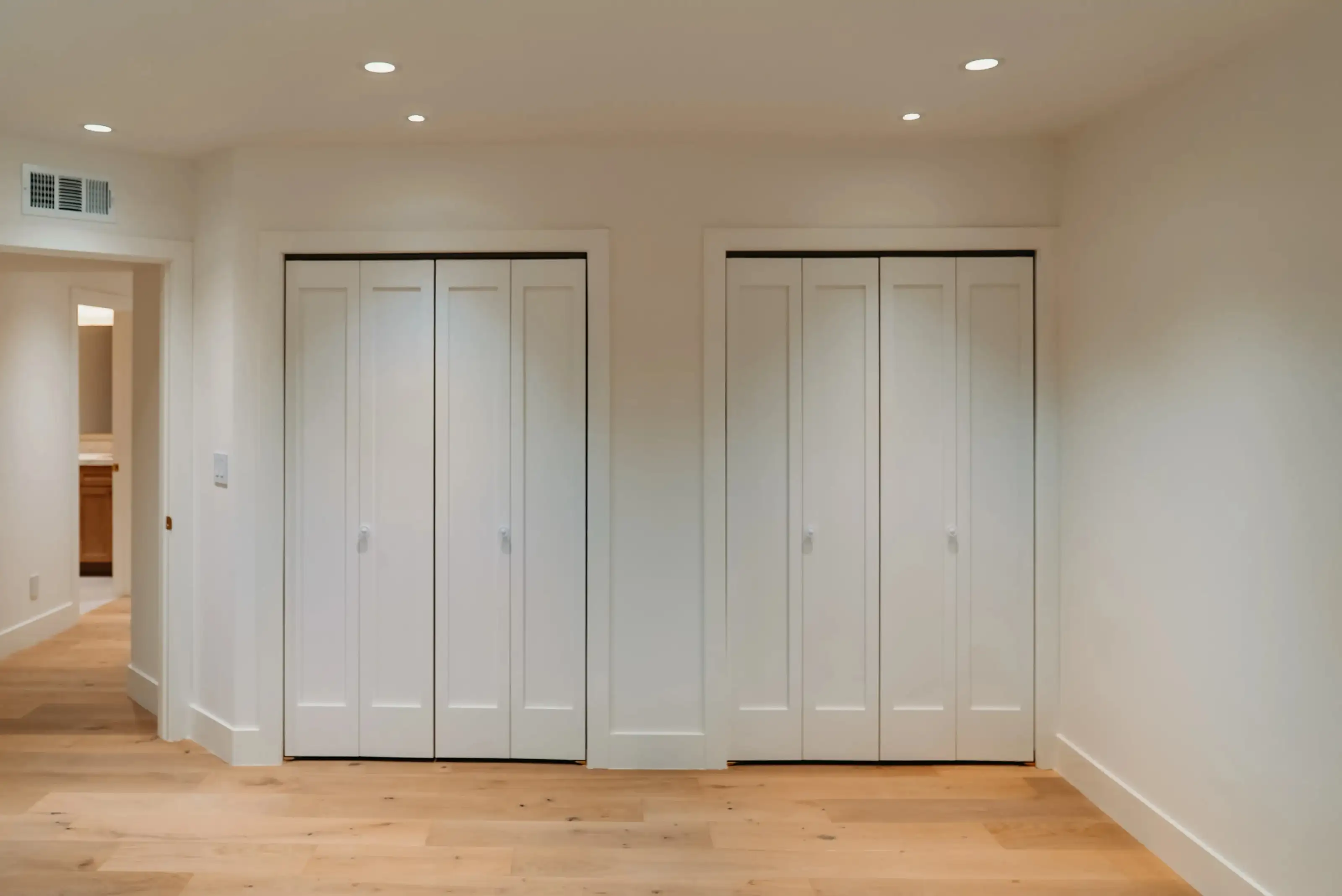 Two white bifold closet doors with shaker panel design installed in an empty room with light wood flooring and recessed