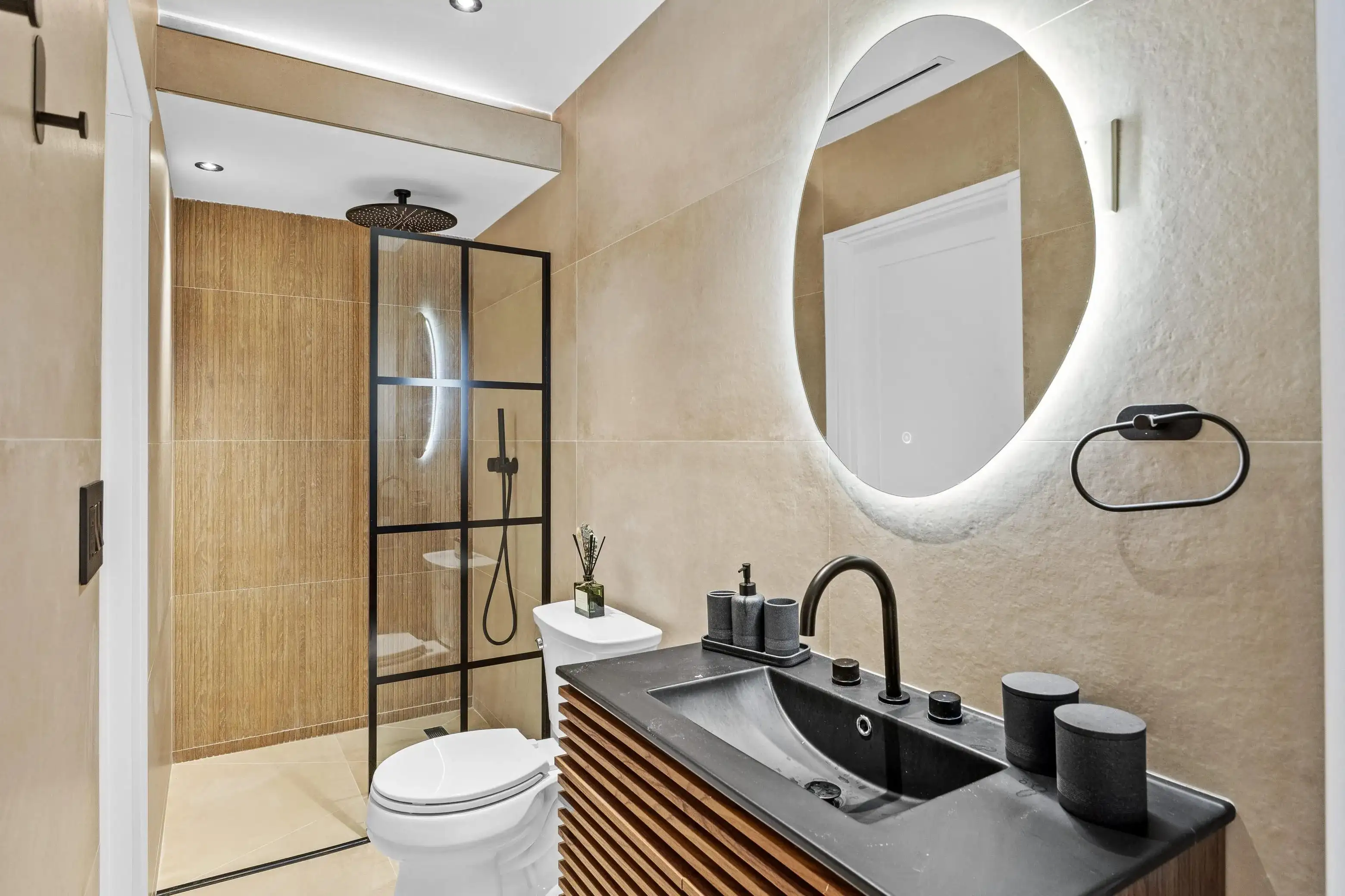 Modern bathroom remodel with black stone sink, wood vanity, backlit round mirror, walk-in shower, and beige tile walls