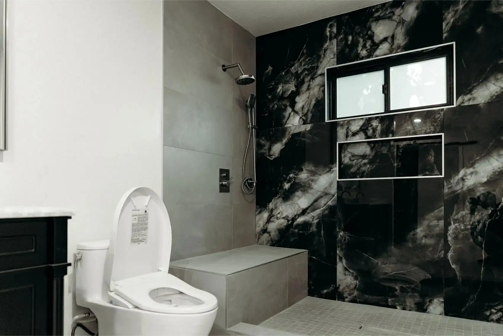 Modern bathroom remodel with black marble shower wall, dual shower system, gray tile bench, and white wall-hung toilet
