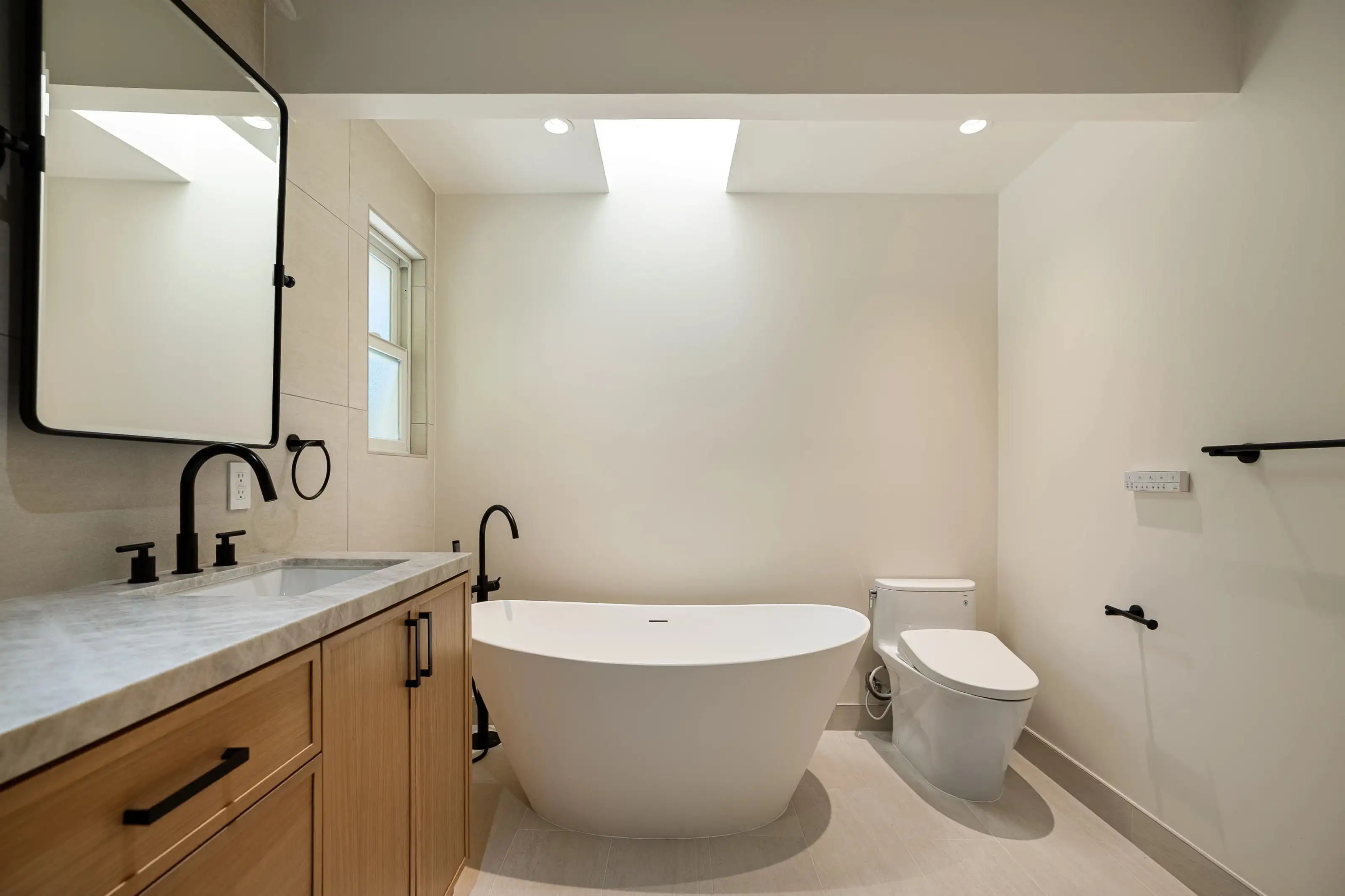 Modern bathroom remodel with freestanding soaking tub, wood vanity, marble countertop, skylight, and matte black fixtures