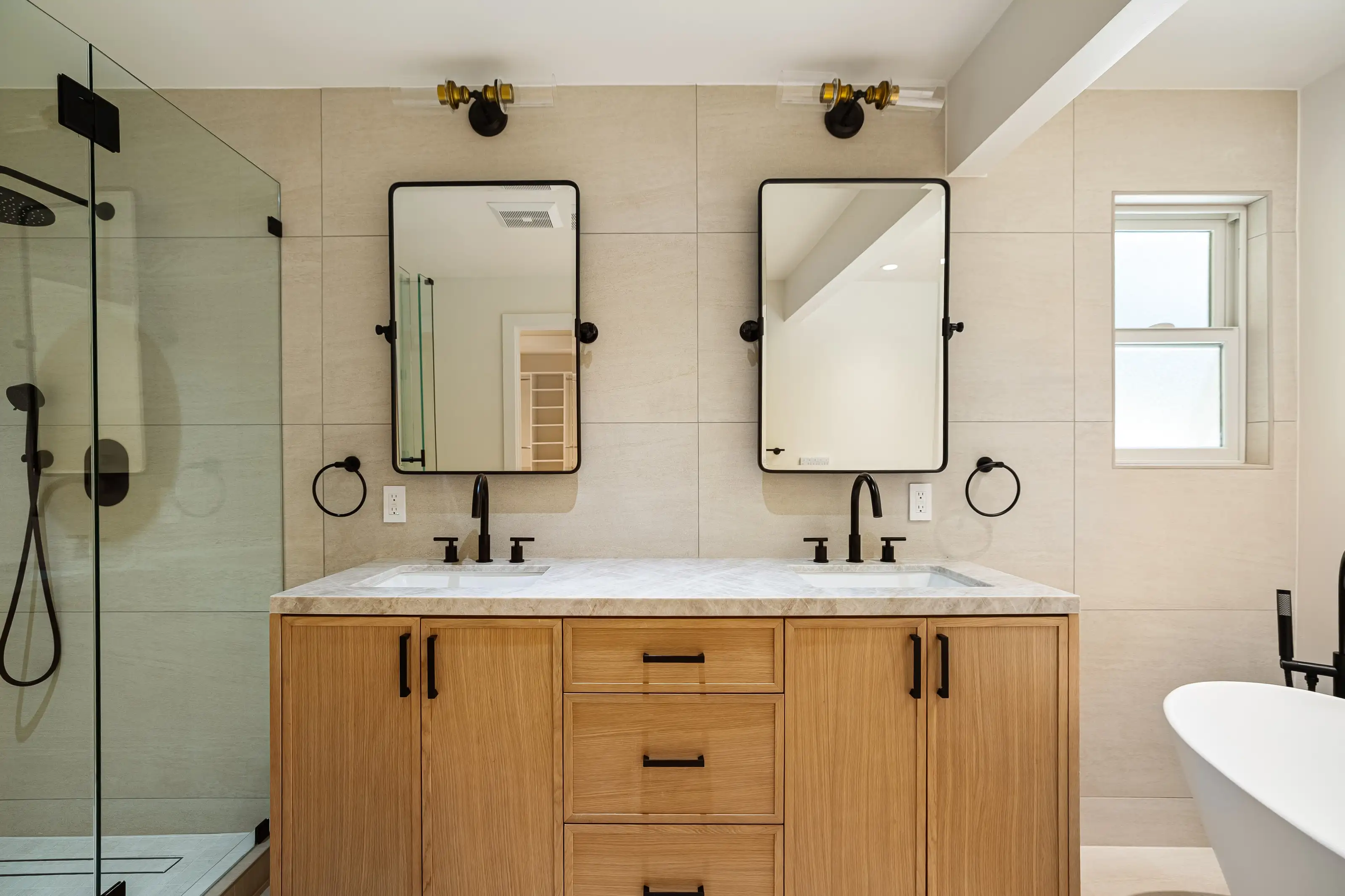 Double sink bathroom vanity with light wood cabinets, marble countertop, black faucets, and pivot mirrors during bathroom
