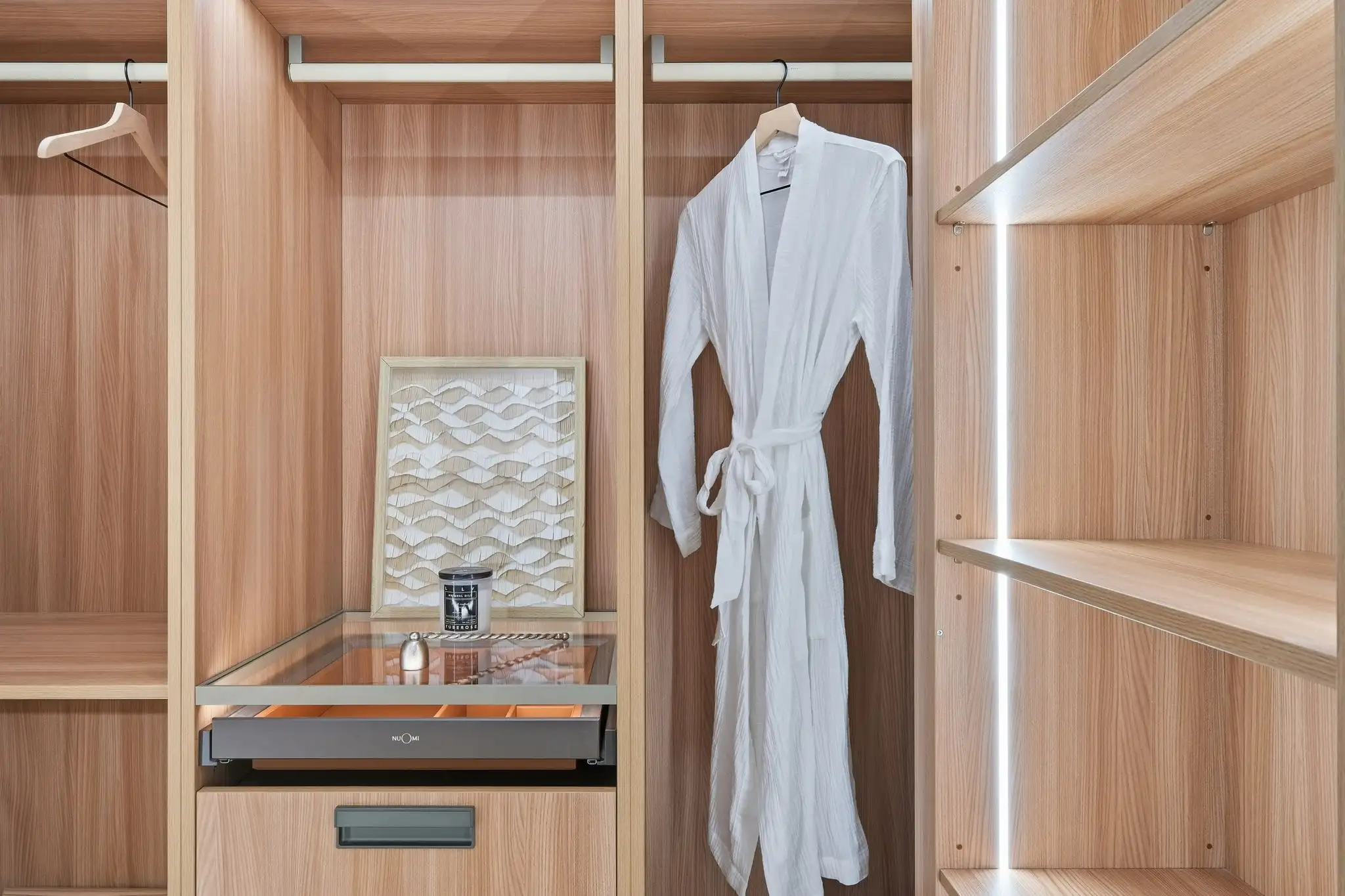 Custom walk-in closet with light wood shelving, white bathrobe on hanger, glass-top jewelry drawer, and built-in lighting