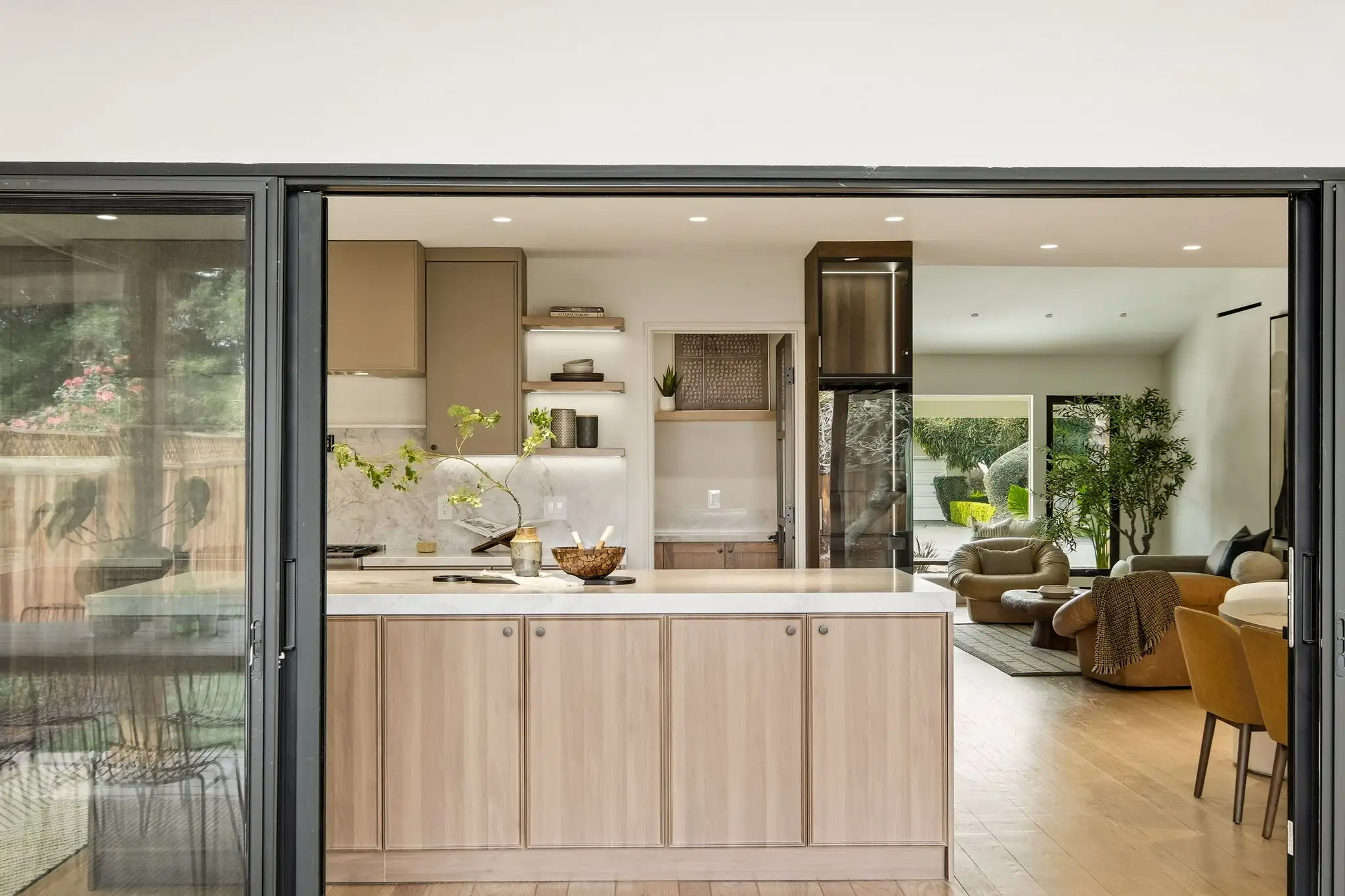 Open-plan kitchen remodel with light wood island, marble backsplash, floating shelves, and folding glass doors to living area