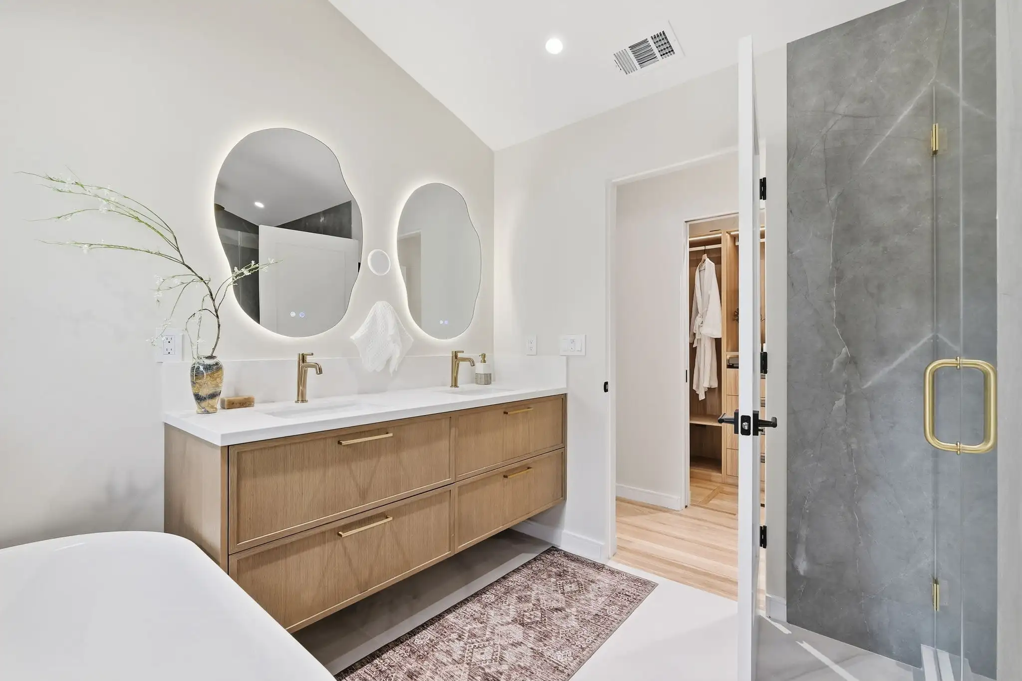 Bathroom remodeling project with floating wood vanity, dual brass faucets, backlit irregular mirrors, and walk-in closet view