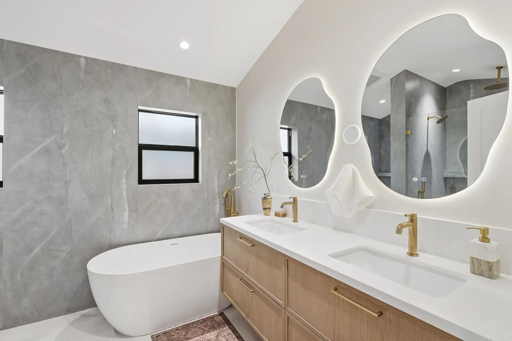 Modern bathroom remodel with freestanding white soaking tub, double wood vanity, gold faucets, and backlit irregular mirror