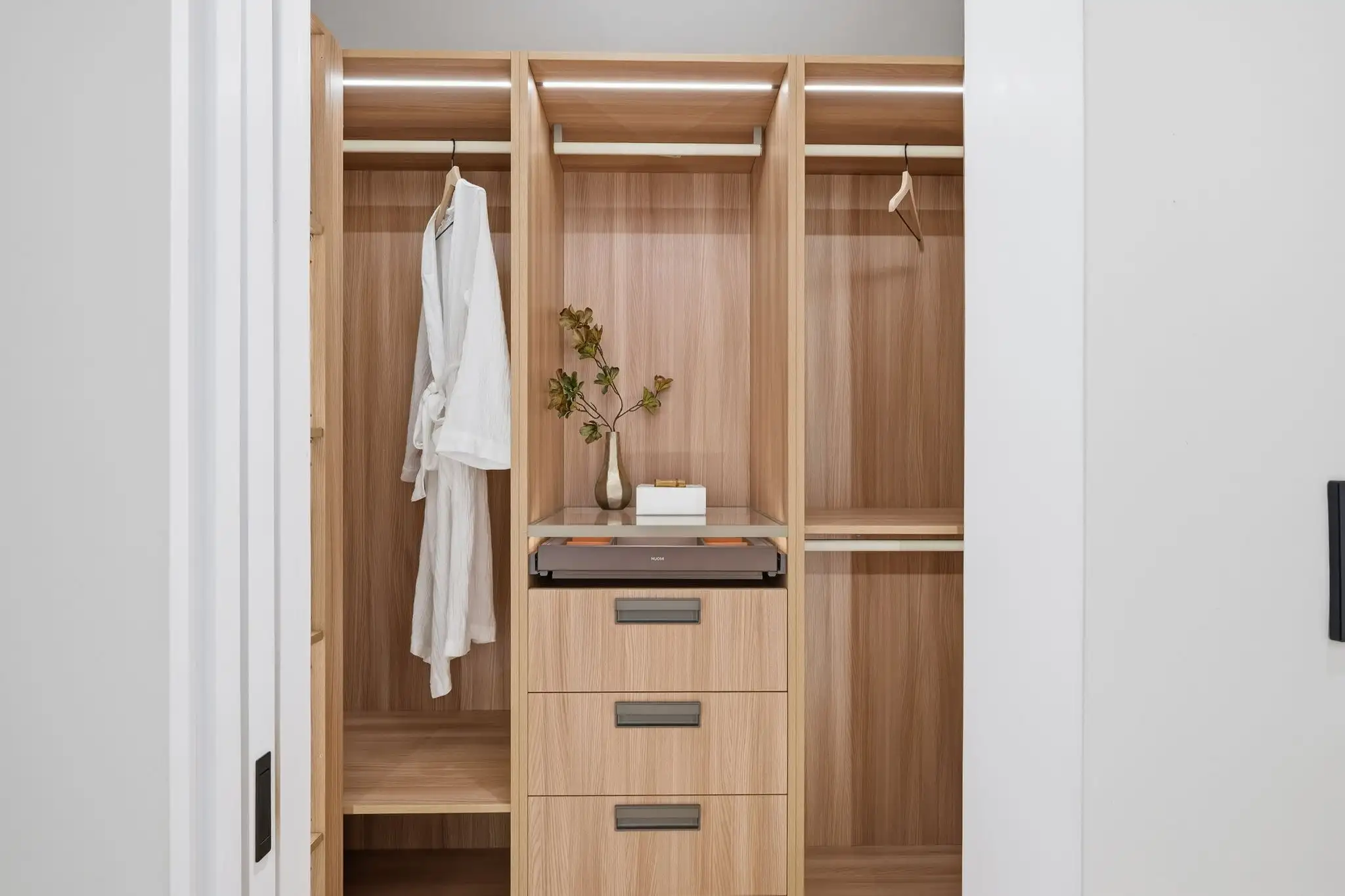 Open walk-in closet with light wood built-in shelving, hanging rods, three drawers, white robe, and decorative plant on shelf