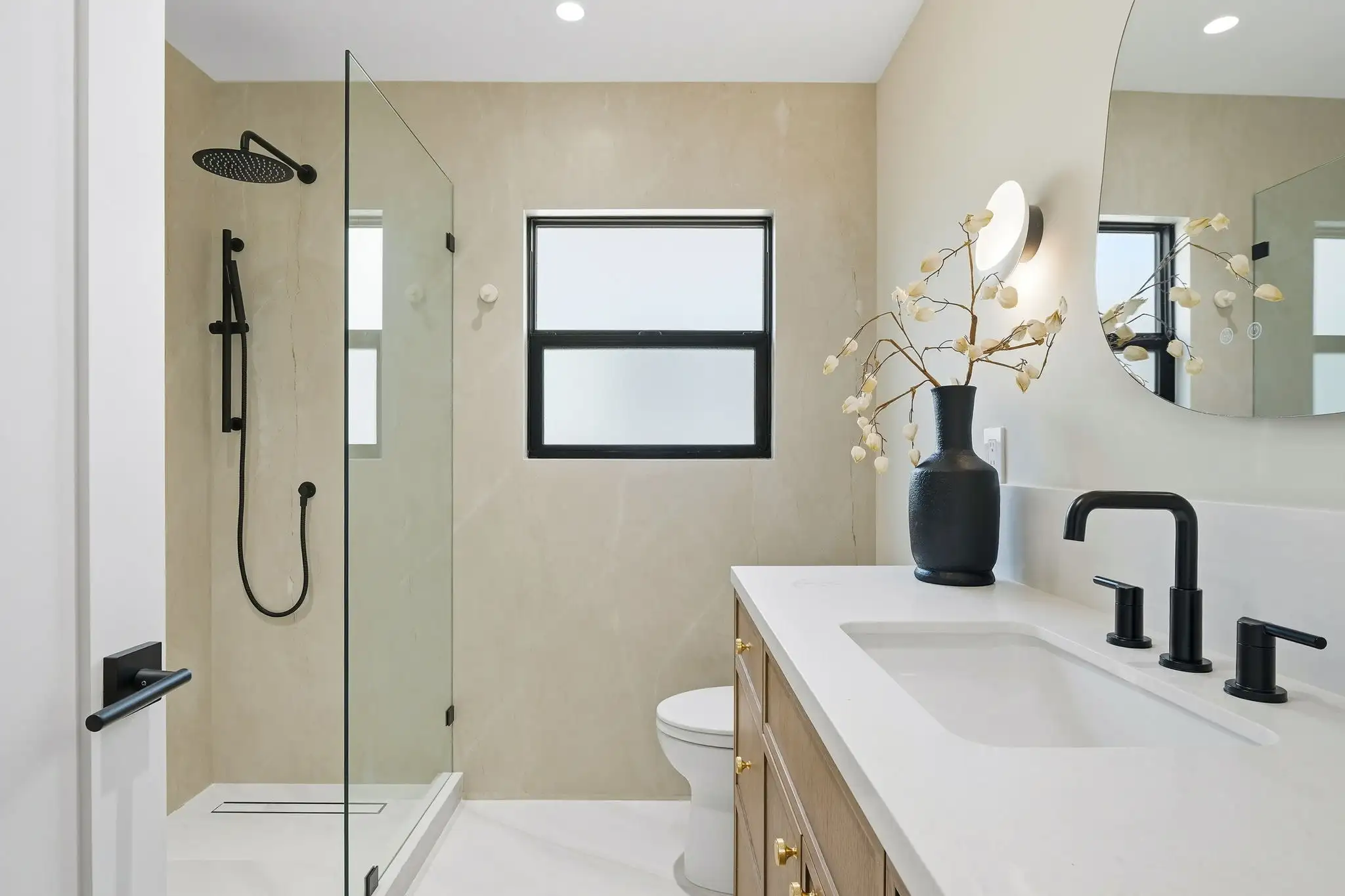 Modern bathroom remodel with glass shower enclosure, wood vanity with gold hardware, black fixtures, and beige stone tile