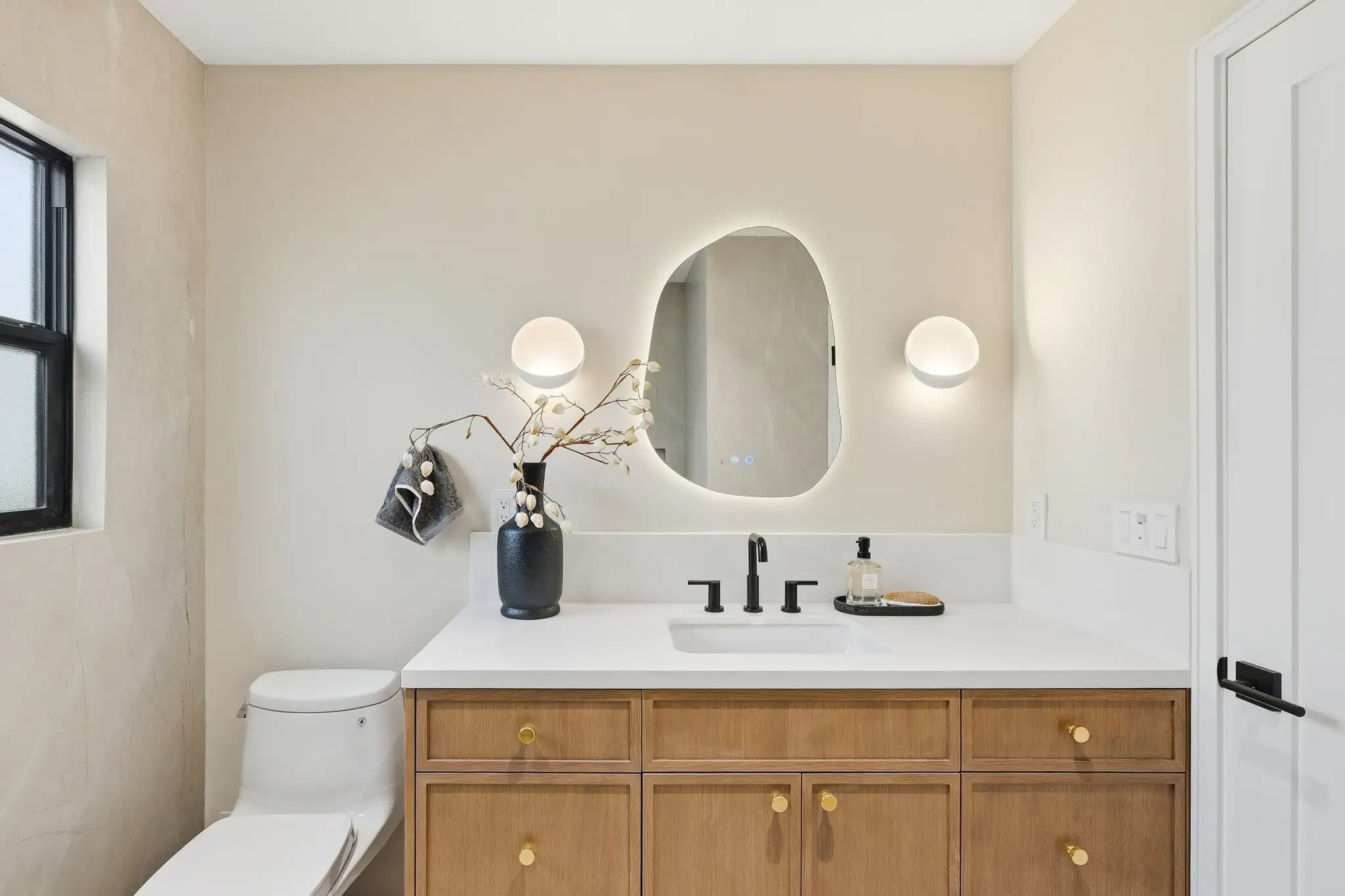 Bathroom remodel with wood vanity, black matte faucet, asymmetric backlit mirror, and globe wall sconces