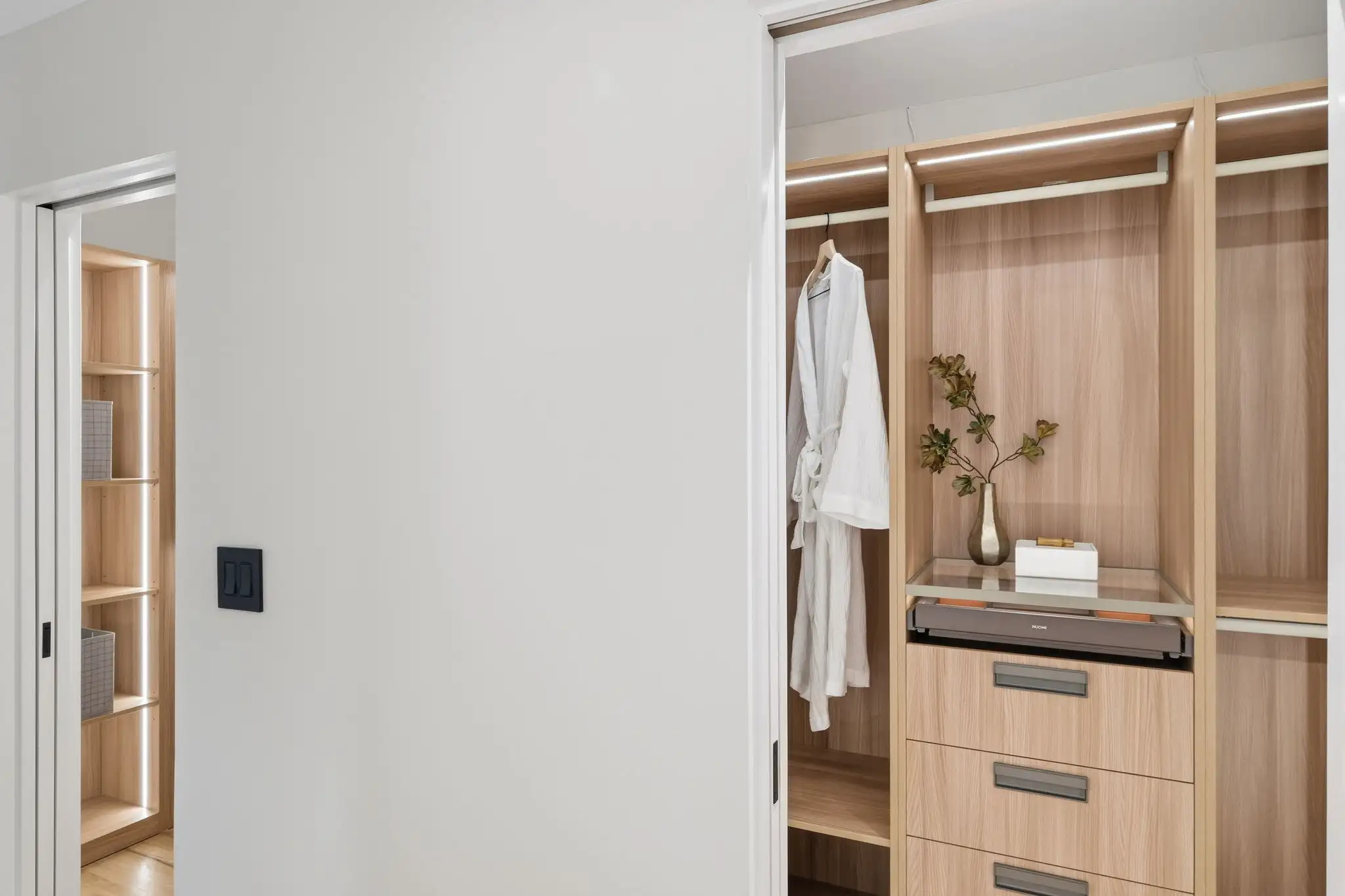 Walk-in closet with white bathrobe hanging on rod, light wood cabinetry, built-in drawers, and decorative vase